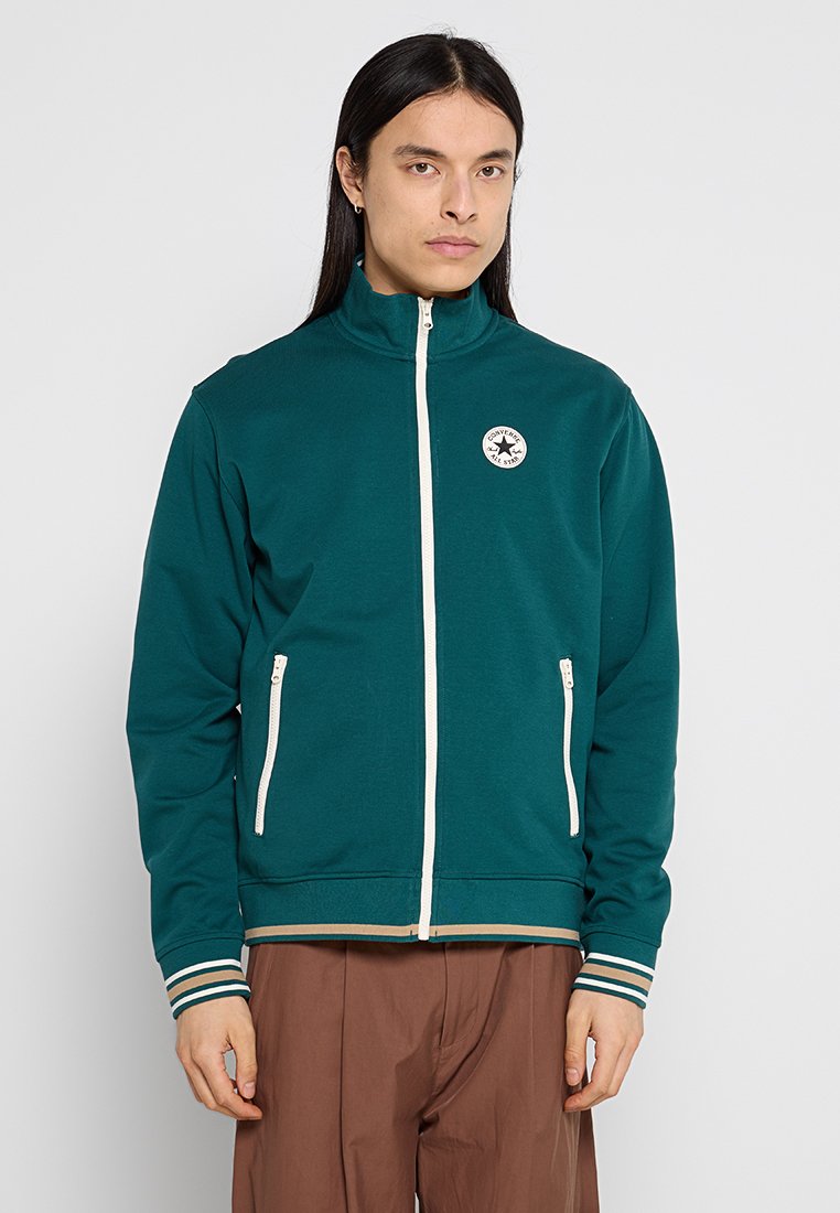 Giacca verde con zip realizzata in materiale morbido, caratterizzata da un collo alto, due tasche frontali, cerniera bianca e polsini a righe. Dettaglio del logo centrale visibile.