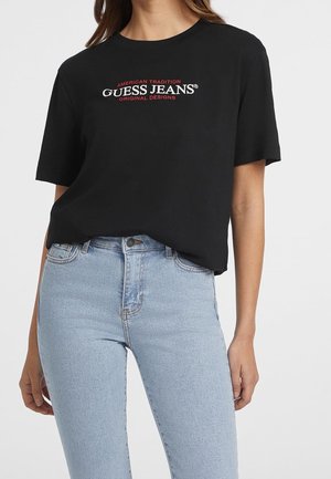 Osoba ubrana w czarną koszulkę Guess Jeans z czerwonym i białym napisem, w połączeniu z jasnoniebieskimi, wysokim stanem dżinsami, obciętymi na wysokości talii.