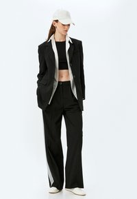 Zwart blazer met een satijnen voering, gecombineerd met zwarte wijde broek met een zilveren zijstreep. Een zwarte crop top en lichte pet maken de look compleet.