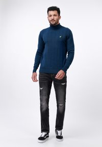 Navyblauw geribbeld turtleneck-trui met lange mouwen, gecombineerd met versleten zwarte jeans en zwart-witte sneakers. Gouden logo-detail zichtbaar.