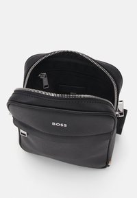 Juodas messenger tipo krepšys su lygiu paviršiumi, sidabrinėmis detalėmis, priekinėmis užtrauktukų kišenėmis ir įspaustu logotipu. Atidarytas, atskleidžia medvilniniu pamušalu vidinę erdvę.