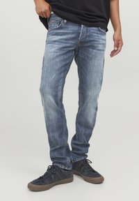 Jeans in denim blu sbiadito con taglio dritto, cuciture visibili e leggermente rovinati; abbinati a sneaker scure.