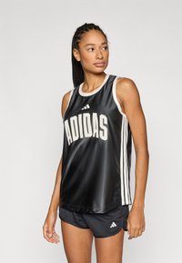 Juoda sportinė liemenė su balta apvadais, priekyje esančiu dideliu užrašu „ADIDAS“, derinamas su juodais sportiniais šortais su trimis dryžiais.