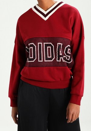 Personne portant un sweat-shirt rouge à col en V avec "ADIDAS" en grandes lettres majuscules sur la poitrine, assorti à un pantalon noir.
