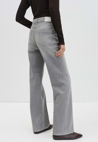 Persoon in hoge taille, wijde grijze jeans met achterzakken, zwart langamige top en puntige donkerenbruine schoenen, staand in een zijaanzicht.