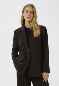 Donna bionda che indossa una giacca di pantaloni scuri marrone e pantaloni coordinati, in piedi con una mano in tasca contro uno sfondo neutro.