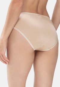 Nainen, joka käyttää béž high-cut saumattomia alushousuja, kuvattuna vyötäröstä alaspäin yksinkertaista taustaa vasten.