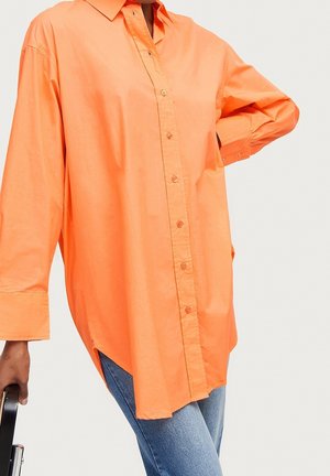 Osoba v oranžové volné košili s dlouhým rukávem na knoflíky a modrých džínách, s jednou rukou v bok a druhou držící černou židli.