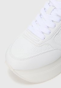 Sneaker bianca caratterizzata da pelle liscia, motivo in rilievo e punta tonda. Include lacci bianchi piatti e una suola spessa e strutturata.