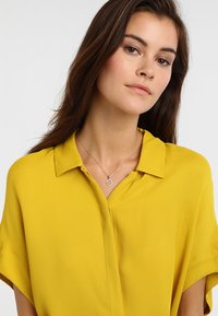 Gul, kortærmet bluse med krave, lavet af letvægtsstof; har en let glans og en afslappet pasform. Sølvvedhæng som accent.