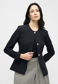 Μαύρο εφαρμοστό σακάκι με στρογγυλή λαιμόκοψη, χρυσά κουμπιά με λουλουδένιο σχέδιο και μακριά μανίκια, συνδυασμένο με καρό φούστα. Λείο υλικό.