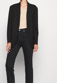 Svart kavaj med strukturerade axlar, en knäppning med en knapp och slag, tillsammans med en beige topp och högmidjade svarta jeans.