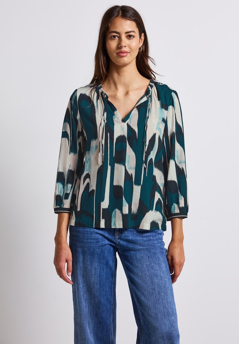 Blouse à motifs teal et beige avec col en V et liens, dotée de manches trois-quarts et d'une coupe décontractée. Assortie avec un pantalon en denim bleu.