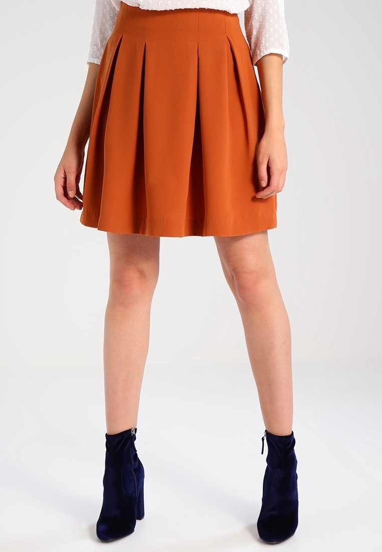 Jupe plissée couleur rouille avec un design évasé, fabriquée en tissu léger, associée à des bottines bleu marine avec un détail zippé.