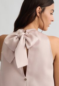 Top senza maniche di colore rosa tenue con un fiocco sul retro, dettaglio a occhiello e chiusura con bottone al collo. Tessuto satinato liscio con una leggera lucentezza.
