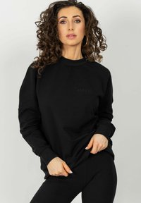 Czarna bluza z ribbowymi akcentami na ramionach, okrągłym dekoltem i wytłoczonym logo. Miękka tekstura z luźnym krojem, w połączeniu z czarnymi legginsami.