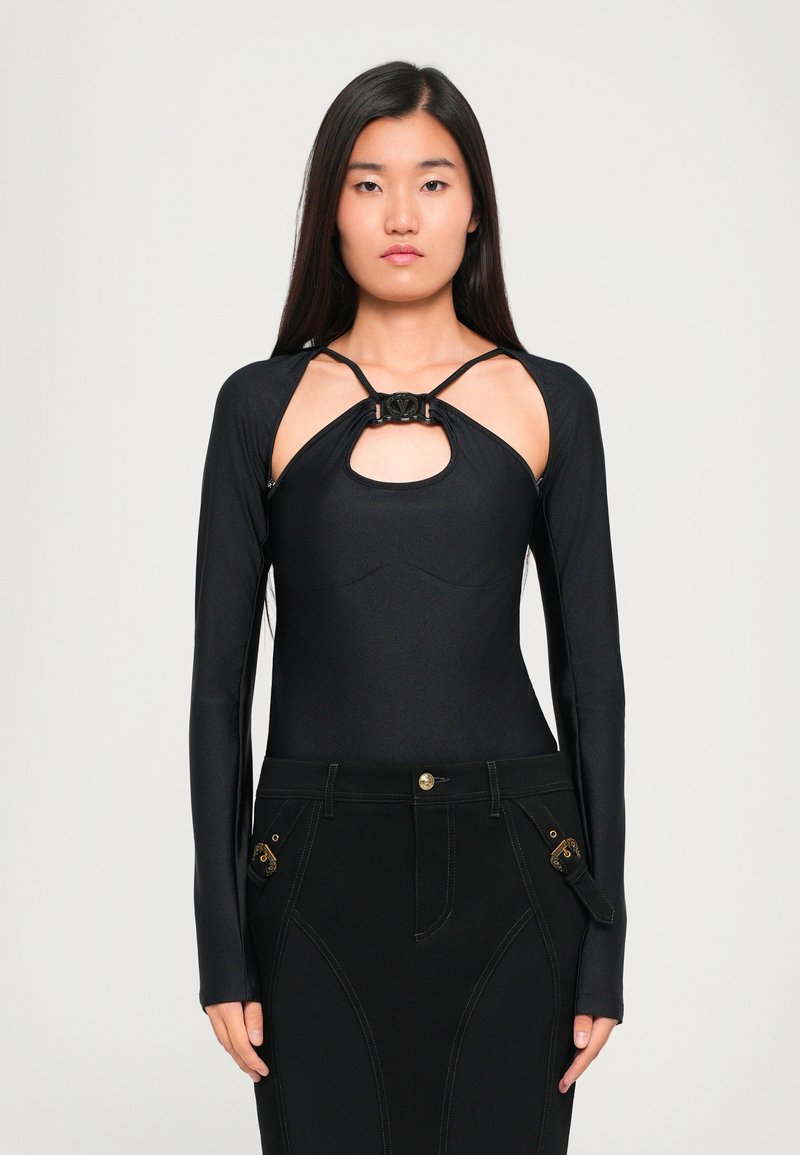 Bodysuit preto de mangas longas com detalhe recortado no peito, tecido texturizado, corte justo, com um nó na gola, combinado com jeans pretos.