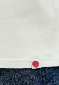 Witte katoenen shirt met een gestructureerde afwerking, voorzien van een rode dotlabel genaaid in de onderzoom, gecombineerd met blauwe denim jeans.