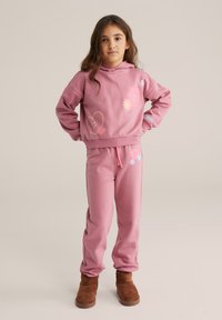 Rózsaszín pulóver és melegítőnadrág szett, színes grafikákkal, húzózsinóros derékkal és puha anyaggal, barna csizmával párosítva.