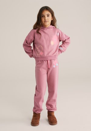 Rosa sweatshirt och sweatpants-set med färgglada grafiska tryck, dragsko i midjan och mjuk tygstruktur, ihop med bruna stövlar.