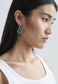 Oorbellen met gouden finish met een grote groene druppelvormige edelsteen omgeven door kleine heldere steentjes, hangend aan een gegraveerde ring.