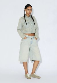 Femme aux cheveux tressés portant une veste en denim claire courte, un short large en denim jusqu'aux genoux et des baskets beige, debout les mains dans les poches.