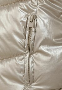 Metallic beige pufferjakke med lynlåslukning, der har en struktureret, skinnende overflade og synlige søm langs sømmen.