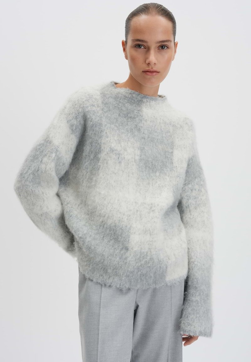 Grau-weißer kariert Pullover aus weichem, flauschigem Material mit lockerer Passform, weiten Ärmeln und Rundhalsausschnitt, kombiniert mit hellgrauen Hosen.