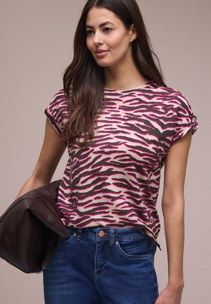 Kvinde med langt mørkt hår iført en pink og sort zebra-mønstret kortærmet top, blå jeans, holder en mørkebrun jakke.