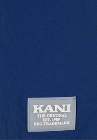 Tamsiai mėlynas audinys su tekstūruotu paviršiumi, savo ruožtu turintis šviesiai pilką stačiakampį etiketę. Etiketėje užrašyta "KANI" ir "EST. 1989" ryškiomis rašmenimis.