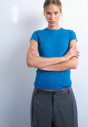 Young woman in blue t-shirt and gray pants stands with arms crossed, looking directly at the camera against a plain light background.