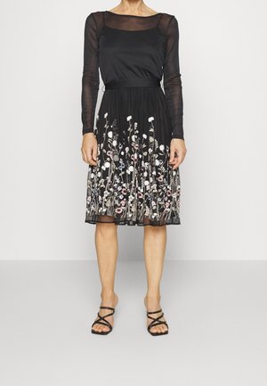 Vrouw draagt een zwarte jurk met doorschijnende lange mouwen en geborduurde bloemenpatronen op de rok, gecombineerd met zwarte sandalen met bandjes.