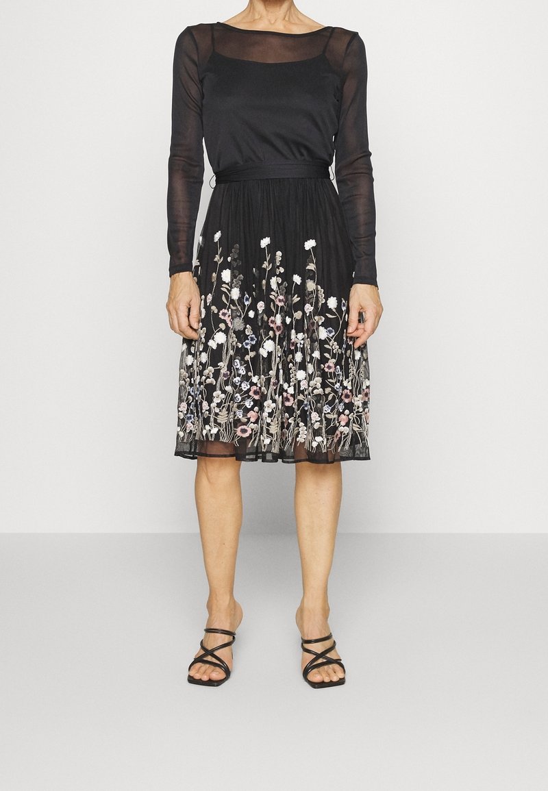 Femme portant une robe noire à manches longues transparentes et motifs floraux brodés sur la jupe, associée à des sandales noires à brides.