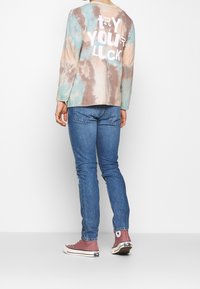 Camiseta de manga larga con diseño tie-dye en colores pastel con texto blanco "INTENTA SUERTE" en la parte trasera, combinada con vaqueros azules y zapatillas rosas.