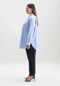 Camicia a righe blu con maniche lunghe e orlo curvo, abbinata a pantaloni neri su misura e sandali neri con tacco alto. Texture del tessuto liscia.