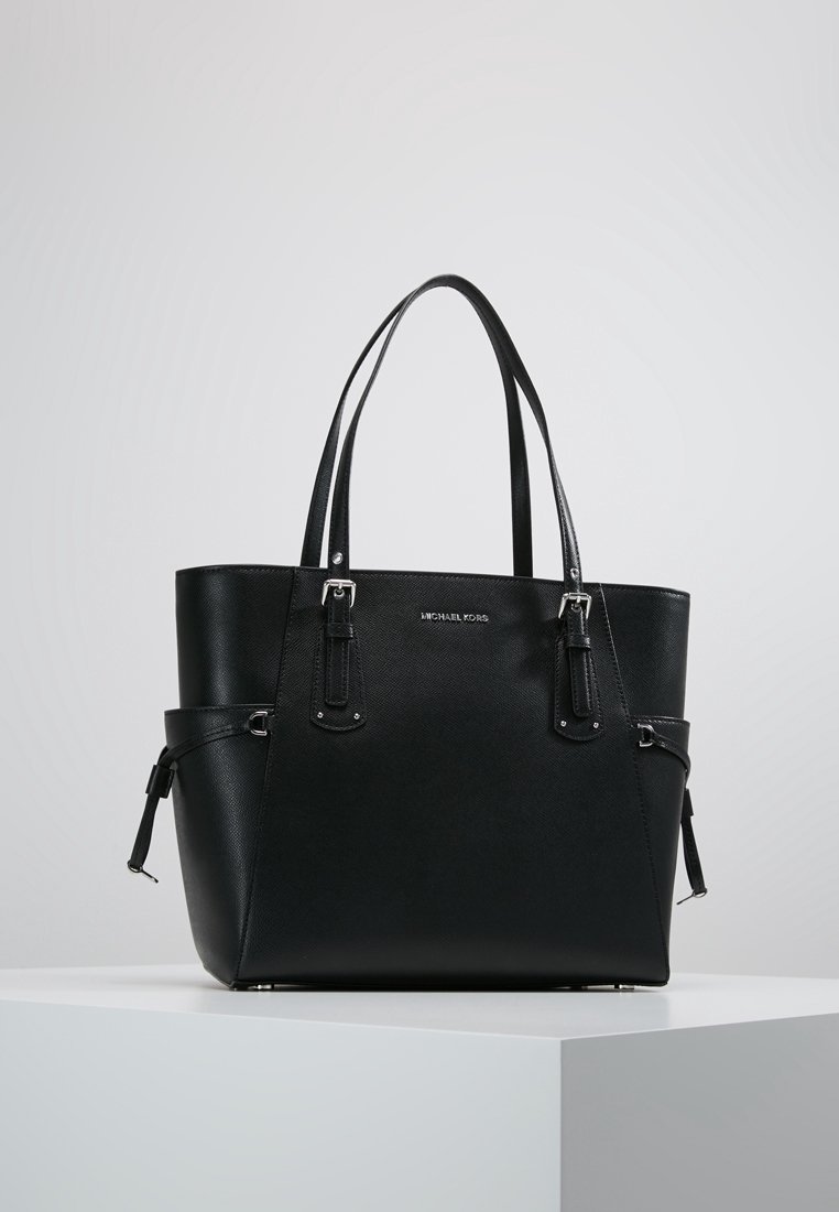 Sac cabas en cuir noir Michael Kors avec doubles bretelles d’épaule et détails de boucles latérales, exposé sur un piédestal blanc sur fond uni.
