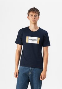 Marineblå bomull t-skjorte med et rektangulært hvitt og gull grafisk merke med teksten "JACK & JONES"; rund hals, korte ermer.