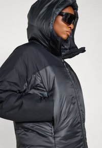 Giacca a vento nera isolata con cappuccio, caratterizzata da una texture liscia e lucida, tasche con zipper e pannelli opachi a contrasto sulle spalle.