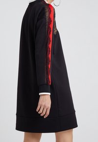 Czarna sukienka dresowa z długimi rękawami, zdobiona czerwonymi i czarnymi koronkami po bokach. Ściągacze na rękawach i biały detal kołnierza.