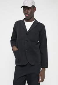 Juodas medvilninis švarkas su įpjauta apykakle, trimis priekinėmis sagomis ir dviem priekinėmis kišenėmis, tekstūruoto paviršiaus. Dėvimas ant balto marškinio.