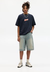 Námořnické oversized tričko s červeným logem, ve spojení s světle modrými džínovými šortkami. Model má na sobě bílé a modré tenisky s kotníkovými ponožkami.
