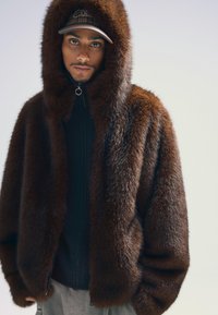 Brun pälsklädd jacka med huva och dragkedja, bärs över en svart stickad tröja. Har en mjuk textur och en oversized passform.