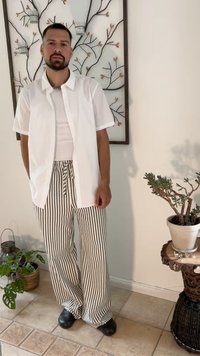 White short-sleeve button-up shirt, white tank top, striped wide-leg pants in black and cream, and black clogs; indoor setting with plants.