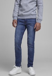 Šedá mikina s malým logem na lemu, kombinovaná s úzkými modrými džíny a bílými teniskami. Jednoduchý, neformální outfit.