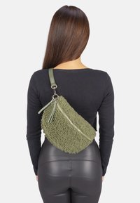 Sac bandoulière en fausse fourrure verte avec une surface texturée, présentant une fermeture éclair diagonale et une sangle en cuir. Sa forme est asymétrique et ergonomique.