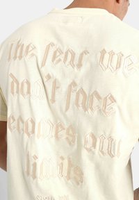 Lys gul bomulds T-shirt med struktureret, præget beige tekst på ryggen med et motiverende citat. Rund halsudskæring, korte ærmer.