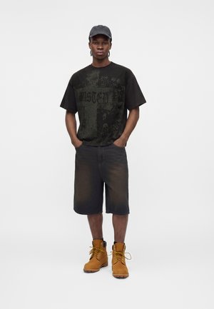 Νέος άνδρας που φοράει μαύρο γραφικό T-shirt, μαύρα τζιν σορτς, ταμπά αρβύλες με κορδόνια και σκουρόχρωμο γκρι καπέλο μπέιζμπολ, στέκεται με τα χέρια στις τσέπες του.