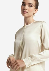 Krämfärgad silkjblus med rund halsringning, långa ärmar och knapppynt vid manschetterna. Slät textur och skräddarsydd passform.