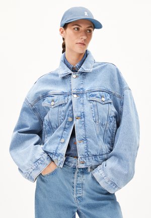 Light blue denim jacket with cropped design, large pockets, and silver hardware. Paired with a matching blue cap.