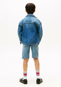 Chaqueta de denim de color azul claro, combinada con pantalones cortos azules y calcetines blancos con rayas rojas y marinas. Calzado negro completa el conjunto.